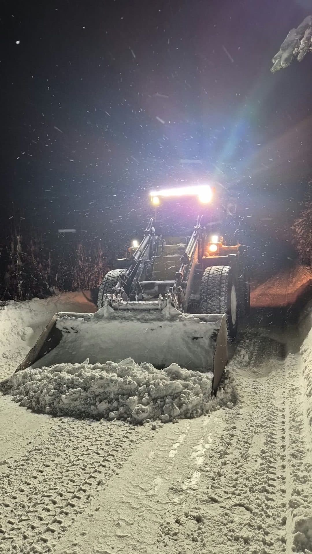 En hjullastare plogar snö på en väg i mörkret, med snöfall i strålkastarljuset
