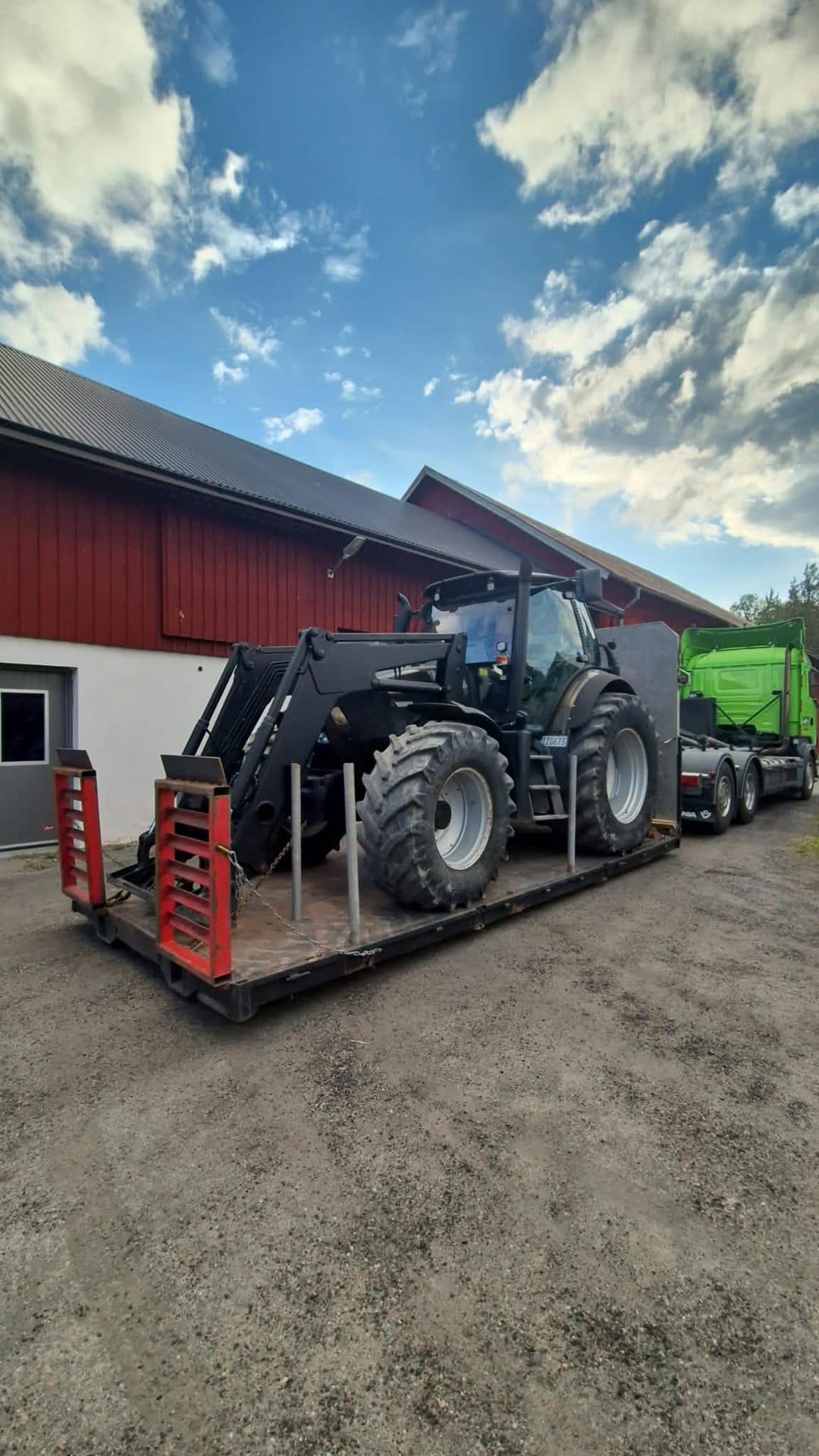 En svart traktor med frontlastare på maskinsläpet, ramperna uppfällda, vid en röd ladugård