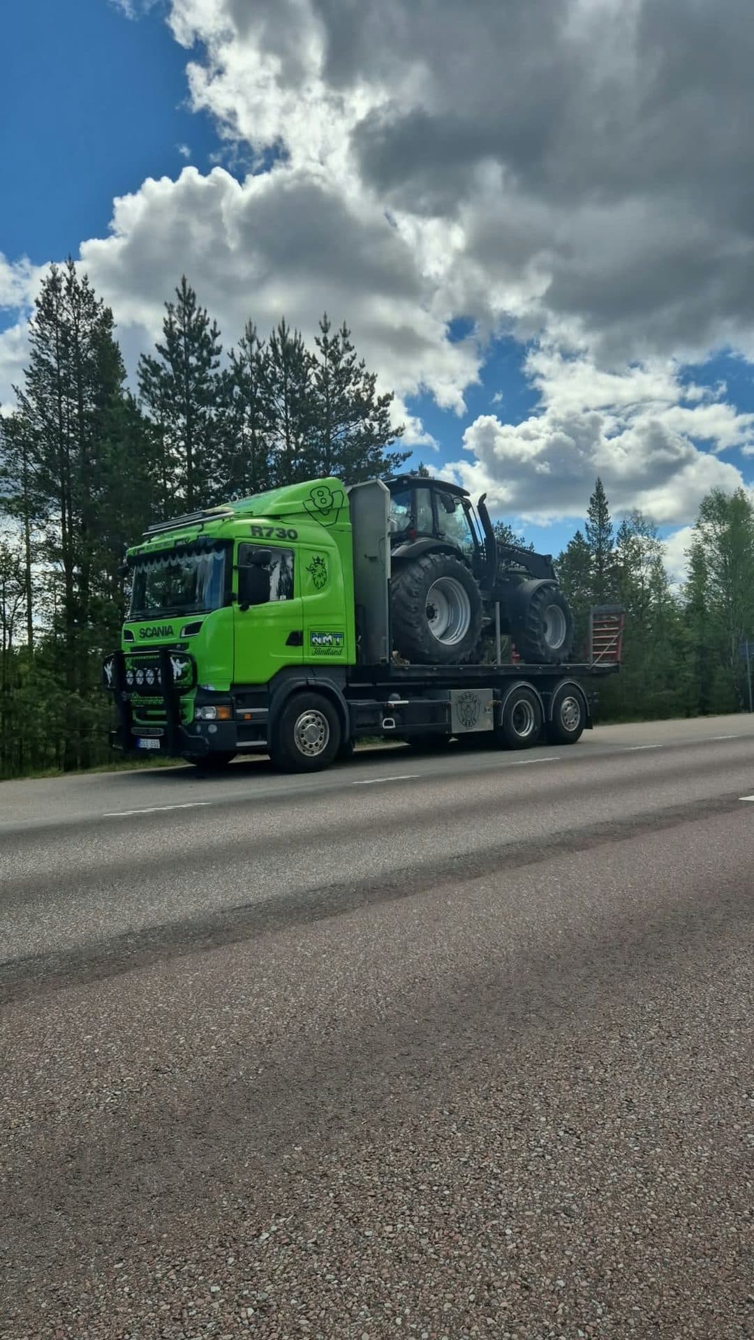 Scanian med en mörk traktor på flaket vid vägkanten, under stora sommarmoln